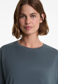 Vrouw met kort golvend bruin haar, gekleed in een donkergrijze ronde hals t-shirt, kijkt recht vooruit met een neutrale uitdrukking tegen een witte achtergrond.