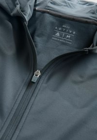 Ciemnoszara kurtka na zamek błyskawiczny o gładkiej fakturze, z srebrnym zamkiem i metką marki na wewnętrznej stronie. Lekka, sportowa konstrukcja.
