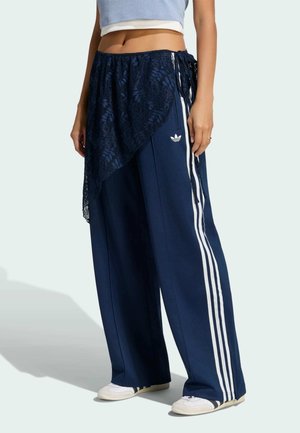 Persona con pantalones sueltos azul marino de Adidas con rayas blancas, zapatillas blancas, un top corto azul claro y una falda cruzada de encaje azul marino transparente.