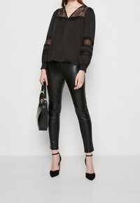 Svart blus med spetsdetaljer på axlarna och i halsringningen, kombinerad med glänsande svarta tajta byxor och en texturerad svart handväska.