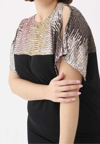 Žena v černých šatech se skvělým multicolor otevřeným ramenním detailem, držící levou ruku pravou rukou.