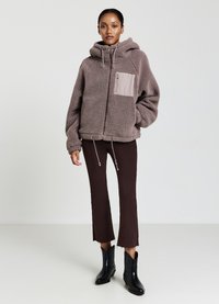 Giacca beige pelosa con cappuccio, zip frontale e tasca laterale; abbinata a pantaloni palazzo marroni e stivaletti neri.