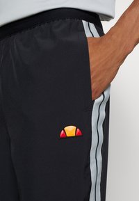 Muž v černých sportovních kalhotách s světle modrými a černými bočními pruhy, ruka v kapse, a malým červeným a oranžovým logem poblíž kapsy.