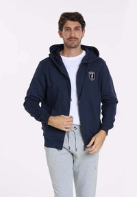 Sweat à capuche zippé bleu marine avec poches avant et un logo sur la poitrine, associé à un pantalon de jogging gris. Tissu doux, coupe décontractée et capuche ajustable.