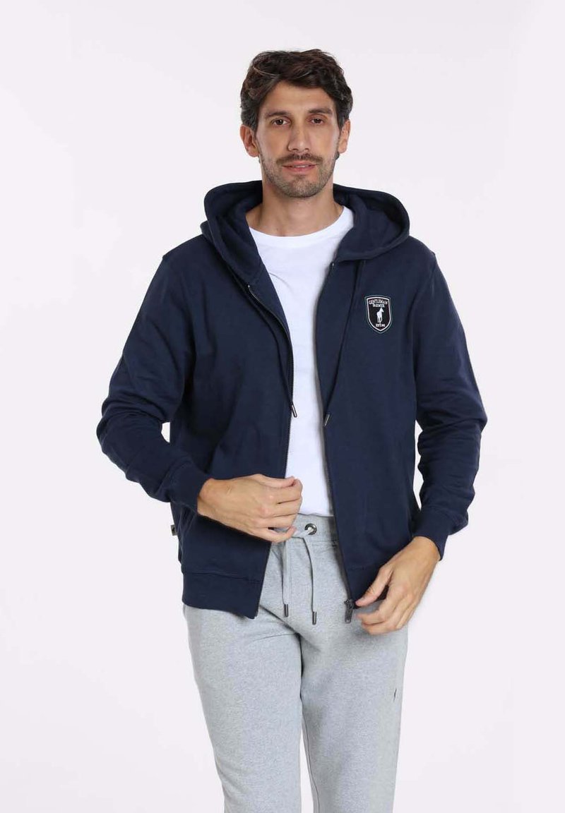 Sweat à capuche zippé bleu marine avec poches avant et un logo sur la poitrine, associé à un pantalon de jogging gris. Tissu doux, coupe décontractée et capuche ajustable.