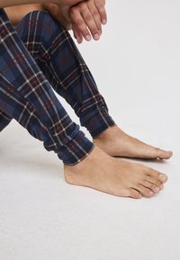 Marinblå rutiga pyjamasbyxor i mjukt tyg, med beige och röda linjer, och avsmalnande muddar. Barfötter synliga i förgrunden på en ljus yta.