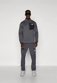 Gray athletic outfit includes a zipped jacket with a chest pocket and gray pants, both with a matte finish. Worn with light gray shoes.