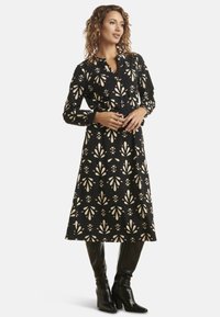 Schwarzes A-Linien-Kleid mit beigem Blumendesign, langen Ärmeln und knöpfbarem Ausschnitt; kombiniert mit eleganten schwarzen kniehohen Stiefeln.