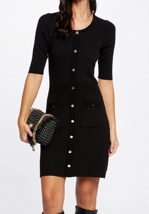 Femme portant une robe noire cintrée boutonnée à manches courtes, tenant un sac à main noir et or à bandoulière chaîne.