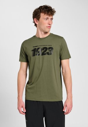 Mężczyzna w oliwkowej koszulce z napisem „hummel SPORTSWEAR SINCE 1923”, w połączeniu z czarnymi spodniami, stoi na tle jednolitego jasnego tła.