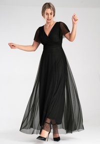 Schwarzes Abendkleid mit einem anliegenden Oberteil, durchsichtigen Flügelärmeln und einem fließenden, transparenten Überzug. Ausgestattet mit einem Wickeldesign und bodenlanger Länge.