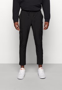 Svarta cargo-byxor med tapered ben, som har två framfickor och elastisk midja. Bärs med vita sneakers. Slät tygkvalitet.