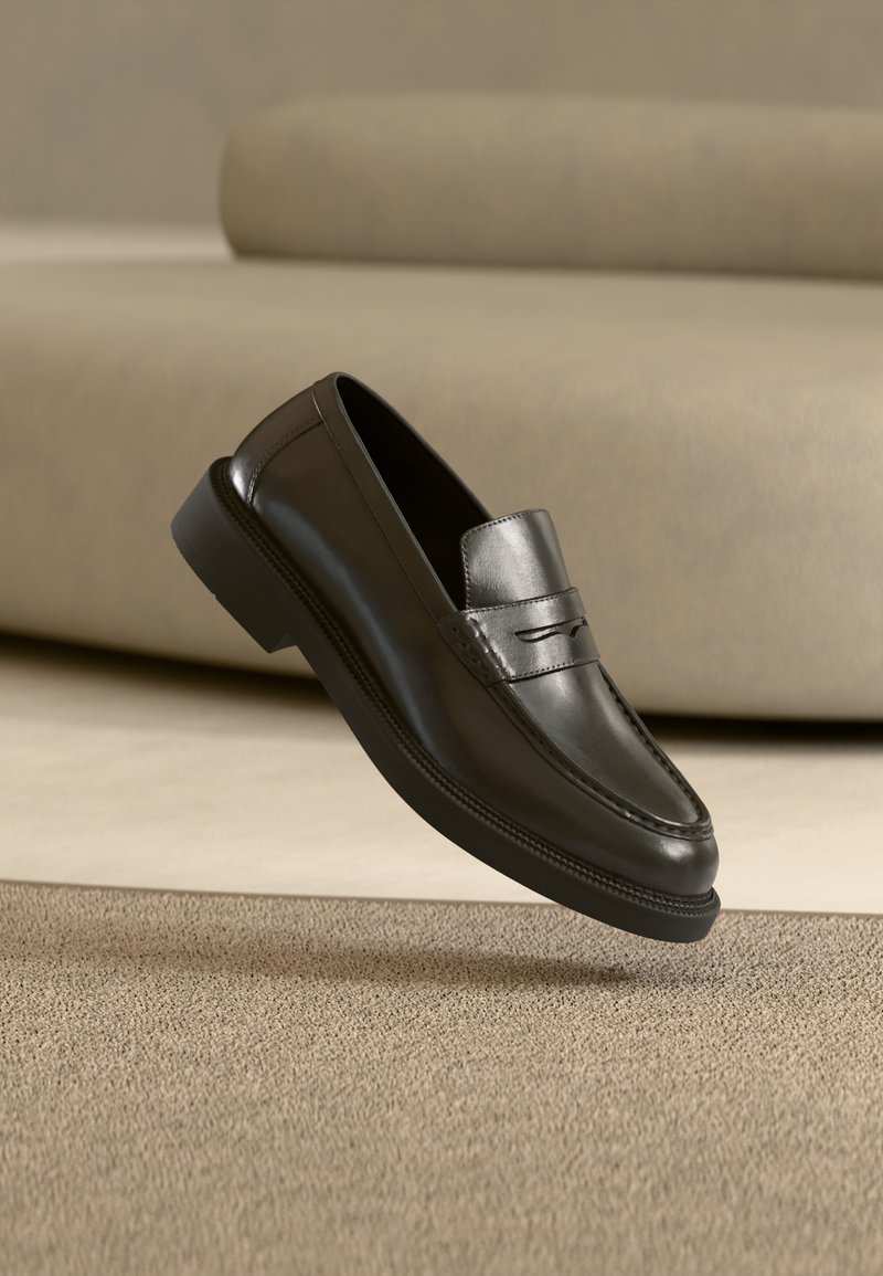 Mocassin penny en cuir noir avec détails cousus flottant au-dessus d'un tapis beige et d'un fond de canapé assorti.