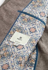 Schmal geschnittene Jacke aus braunem Textil mit blauem Futter, das geometrische Muster in Blau, Orange und Creme aufweist, sowie einem Etikett.