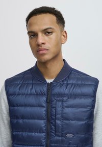 Mlad čovjek s kratkom kovrčavom kosom nosi tamnoplavi quilty prsluk preko svijetlosivog dugog rukava, u neutralnoj svijetloj pozadini.