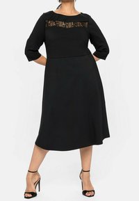 Robe noire à manches trois-quarts, avec un panneau en dentelle transparente au niveau du col. Tissu lisse, forme évasée, et ourlet jusqu'au genou.