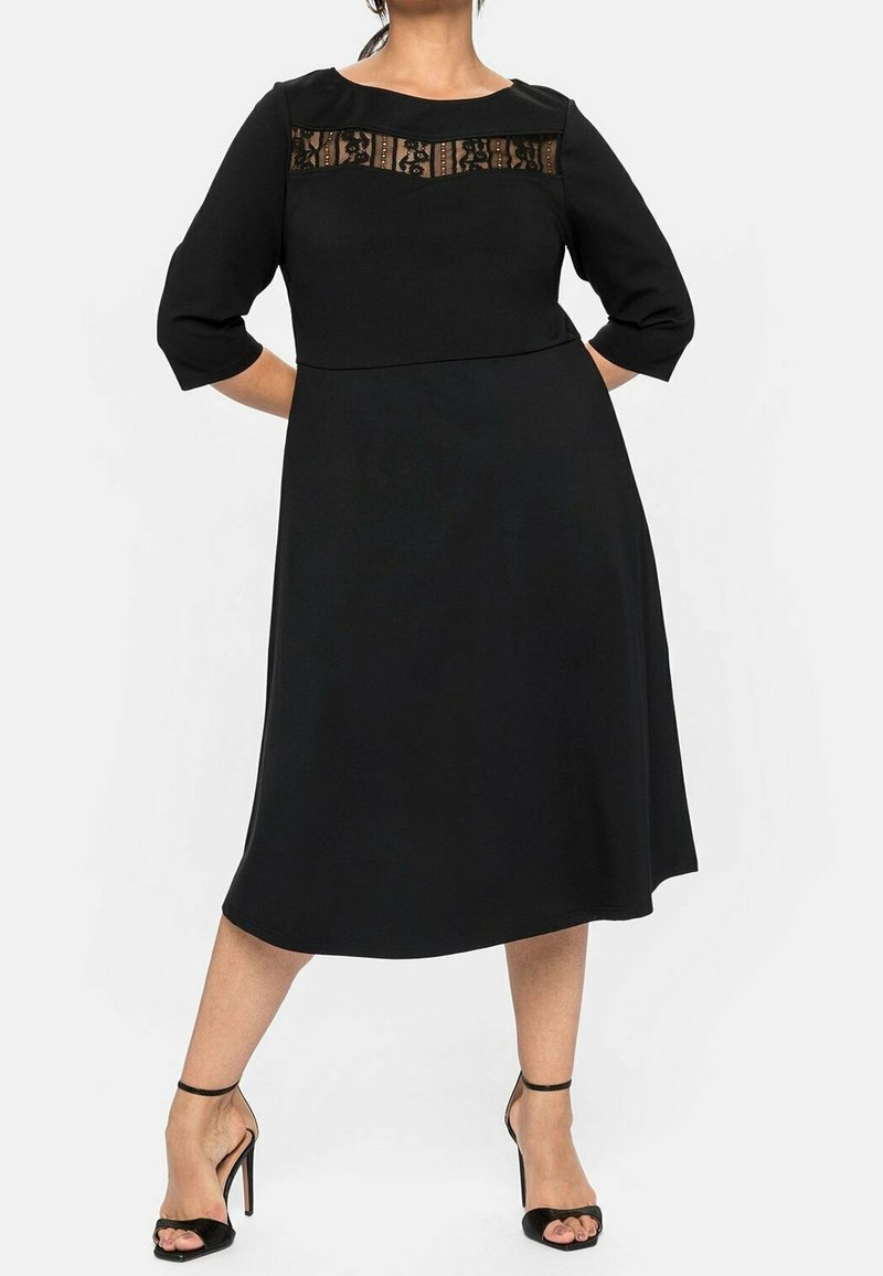 Robe noire à manches trois-quarts, avec un panneau en dentelle transparente au niveau du col. Tissu lisse, forme évasée, et ourlet jusqu'au genou.