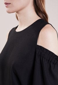 Svart topp med bred halsringning, kallskulderdesign och sammangränsade ärmdekorationer, tillverkad av ett mjukt, lätt tyg.