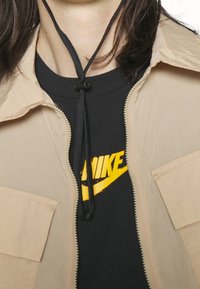 Giacca beige con zip e finitura testurizzata, con un laccetto nero al collo, indossata sopra una t-shirt nera con un logo giallo.