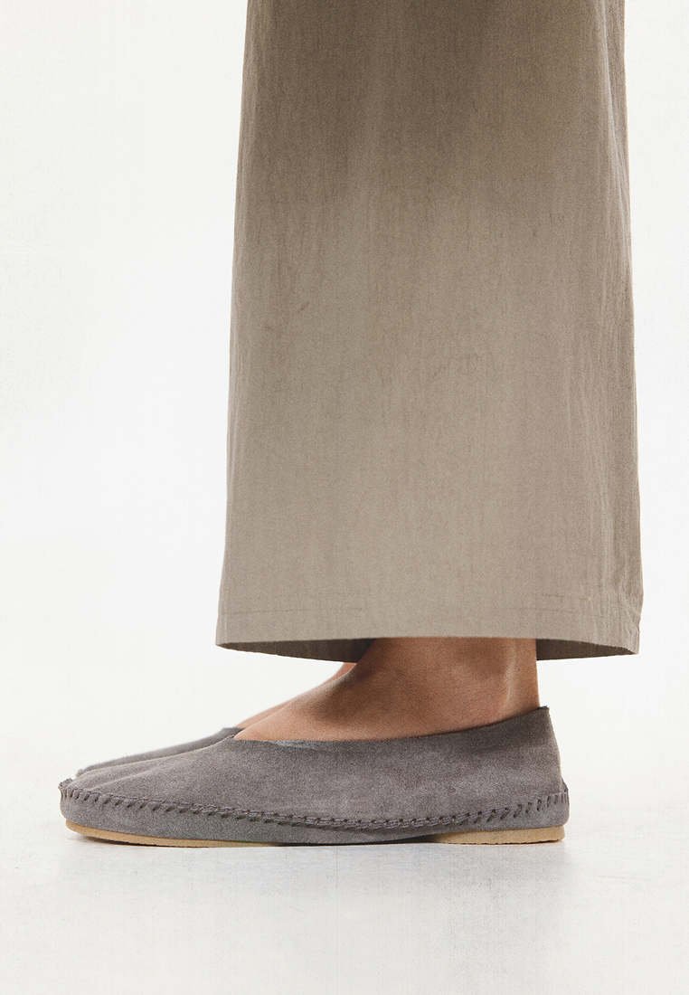 Pieds chaussés de chaussures gris suède sans lacets avec détails de couture, associés à un pantalon beige à jambes larges, vus de profil sur un fond clair.
