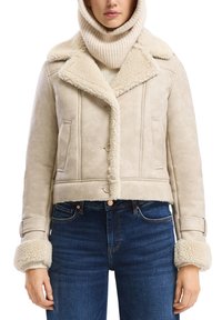 Vrouw die een beige schapenvachtjasje met brede kraag en manchetten draagt, gecombineerd met blauwe jeans en een crèmekleurige coltrui.