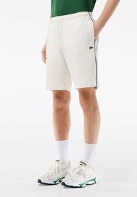 Shorts de sport blancs avec des bandes vertes sur les côtés, taille élastique et logo Lacoste. Portés avec des chaussettes blanches et des baskets avec des accents verts.