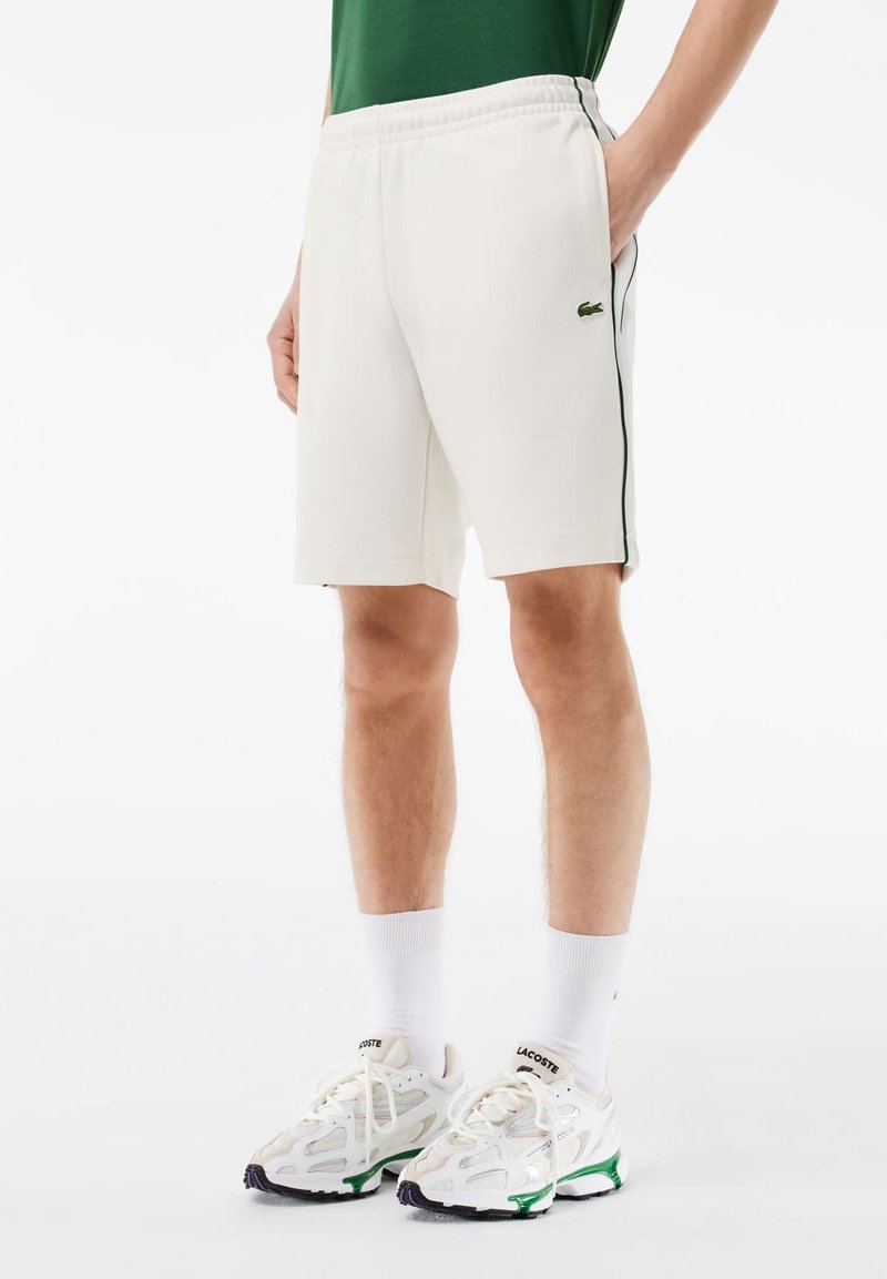 Shorts de sport blancs avec des bandes vertes sur les côtés, taille élastique et logo Lacoste. Portés avec des chaussettes blanches et des baskets avec des accents verts.