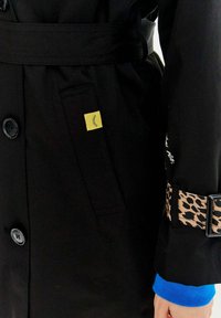 Čierny trenčkot so žltou nášivkou, so opaskom s leopardím vzorom a modrými manžetami. Obsahuje zapínanie na gombíky a detaily vrecka.