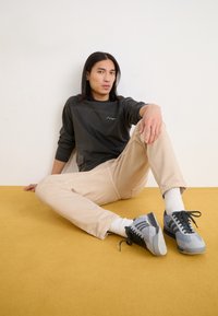 Sweatshirt vert foncé avec un motif texturé, pantalon beige et baskets grises avec des accents noirs et des chaussettes blanches, assis sur une surface jaune.