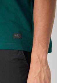 Zeleni pamučni T-shirt kratkih rukava, s teksturiranom crnom oznakom s bijelim logotipom blizu rubnog dijela. Crne kratke hlače djelomično vidljive.