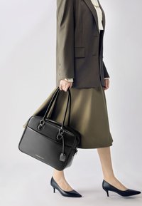 Sort læderhåndtaske med to håndtag, rektangulær form, guldfarvede detaljer og et mærke. Båret af en person i et brunt jakkesæt og hæle.