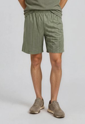 Homme portant un short rayé vert olive, une chemise assortie et des baskets beige, debout les mains dans le dos sur un fond uni.
