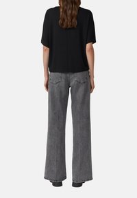 Kobieta z falującymi brązowymi włosami, ubrana w czarny krótki top i szare dżinsy o wysokim stanie i szerokich nogawkach, stoi tyłem na jednolitym tle.