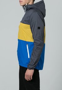Giacca a blocchi di colore con una parte superiore a scacchi neri, una sezione centrale gialla e una parte inferiore blu. Include un cappuccio e una tasca frontale con zip.