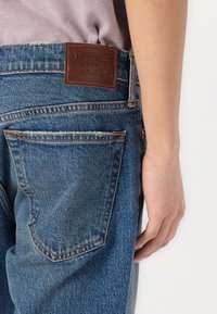 Vaqueros de denim azul oscuro con un acabado texturizado. Presentan una etiqueta de marca de cuero marrón y costuras en contraste. Bolsillo visible en el lateral.