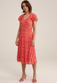 Floral-patterned pink dress with short ruffled sleeves, V-neckline, tied waist, and a flowing skirt. Paired with red strappy sandals.
