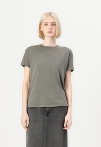 Olivgrünes T-Shirt aus weichem Baumwollstoff, mit kurzen Ärmeln und Rundhalsausschnitt, getragen zu einem grauen Jeansrock. Leicht strukturierte Oberfläche.