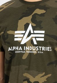 Πράσινο καμουφλάζ T-shirt από βαμβάκι. Διαθέτει λευκό λογότυπο με στυλιζαρισμένο κείμενο "ALPHA INDUSTRIES" και λεπτομέρειες θέσης σε έντονα γράμματα από κάτω.