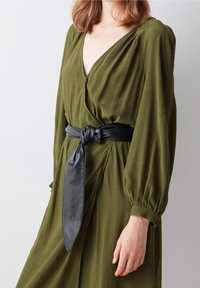 Femme portant une robe à manches longues vert olive avec une large ceinture noire nouée à la taille, debout devant un fond uni.