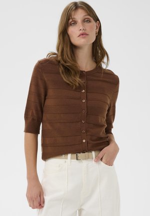 Žena oblečená v hnedom krátkom rukáve s gombíkmi a bielych nohaviciach s vysokým pásom, so svetlobežovým opaskom, stojí s jednou rukou v vrecku.