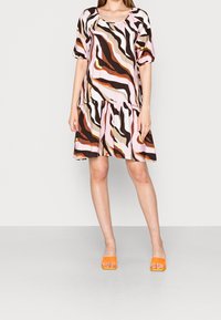 Mujer vistiendo un vestido corto y suelto con patrones abstractos en tonos rosa, marrón, naranja y negro, combinado con unos tacones destalonados de color naranja brillante.