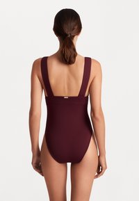 Fato de banho integral bordeaux com alças largas e costas baixas. Textura suave com um brilho subtil e um detalhe metálico do logotipo na cintura.