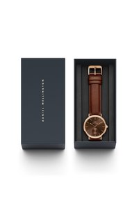 Brown leather strap watch with a dark brown face, gold-tone accents, and a round case. Presented in a dark blue box with black interior.