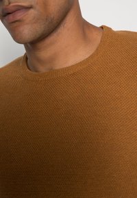 Närbild av en mans nedre ansikte och nacke som bär en strukturerad brun rundhalsad tröja mot en enfärgad bakgrund.