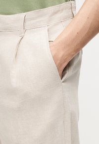 Χέρι που είναι τοποθετημένο στην μπροστινή τσέπη μπεζ παντελονιού που φοριέται με πράσινο πουκάμισο, δείχνοντας την υφή του υφάσματος και τις λεπτομέρειες ραφής.