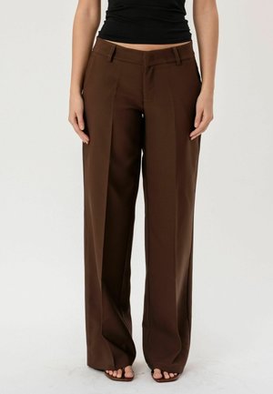 Kobieta w brązowych, szerokich spodniach z wysokim stanem i czarnym, bezrękawnym topie, stojąca z rękami swobodnie opuszczonymi wzdłuż ciała na jednolitym tle.
