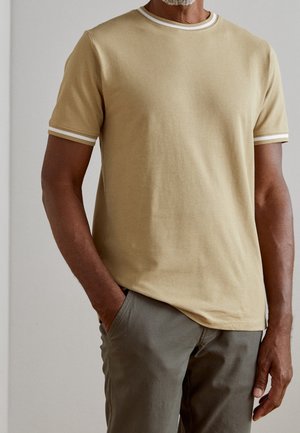 T-shirt a maniche corte, di un beige chiaro con strisce bianche di accento sul colletto e sulle maniche, abbinato a pantaloni verde oliva. Tessuto dalla texture liscia.