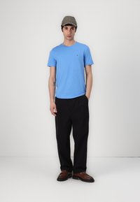 Hellblaues Baumwoll-T-Shirt mit Rundhalsausschnitt und kurzen Ärmeln; kombiniert mit schwarzen, lässig sitzenden Hosen und braunen Slippers. Akzent-Logo auf dem Shirt.