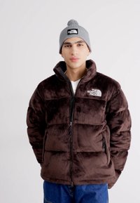 Brūna mākslīgās kažokādas puffer jaka ar rāvējslēdzēju priekšā un sānu kabatām; pelēks adīts beanie ar logo; modele stāv, rokas atvieglinātas pie sāniem.