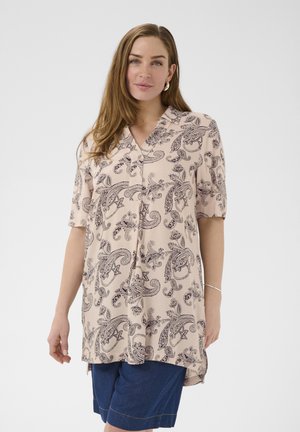 Žena nosi svetloružovú blúzku s paisley vzorom a krátkymi rukávmi a modré riflové šortky, stojaca na jednofarebnom pozadí.